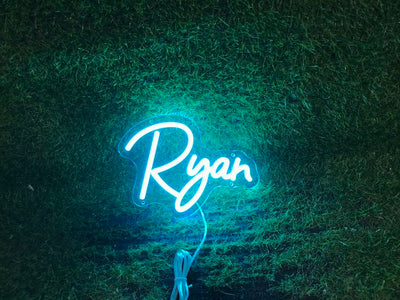 Ryan Name Neon sign in stylish script, mounted on a clear acrylic panel and radiating vibrant light against a modern interior wall - Neon.Outshine 